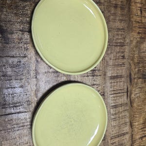 May include: Two pale yellow ceramic plates with a slightly irregular shape. The plates have a glossy finish and a raised rim. They are displayed on a rustic, dark wood surface, creating a contrast in textures and colors. The plates are suitable for serving food.