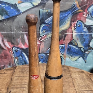 May include: Two vintage wooden exercise clubs standing upright. The left club features a red Sportcraft label. The right club is taller, has a black painted ball top, two black horizontal bands, and the text Wilson Jr. stamped on the wood.