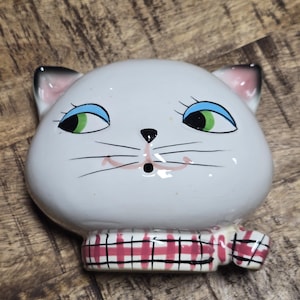 May include: A white ceramic cat head with blue eyes, green irises, and a pink mouth. The cat has black whiskers and a red and black plaid scarf. The ears are black-tipped with pink interiors. A decorative item.