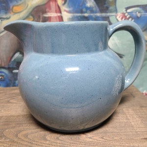 May include: A light blue ceramic pitcher with a rounded body and a handle. The pitcher has a speckled glaze.