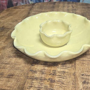 May include: A pale yellow ceramic serving dish with a ruffled edge and a matching small bowl in the center. The dish is set on a rustic wooden table, creating a charming, vintage aesthetic. Ideal for serving snacks or dips.