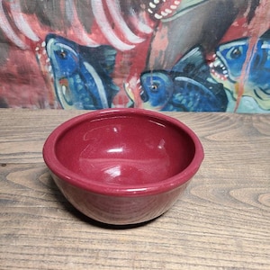 May include: A deep red ceramic bowl with a smooth, glossy finish. The bowl is empty and sits on a wooden surface.