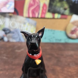 May include: A glossy black dog figurine with a red collar and a yellow tag. The dog is sitting upright with its ears perked up. The figurine is on a brown surface.