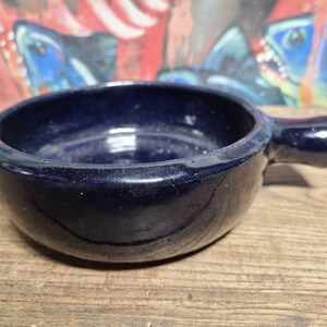May include: A dark blue ceramic ramekin with a handle. The ramekin is glazed and has a smooth, rounded interior.