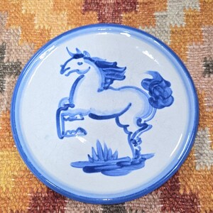 May include: A white ceramic plate with a blue rim and a blue horse illustration. The horse is depicted in a running pose. The plate is round and sits on a colorful patterned surface.