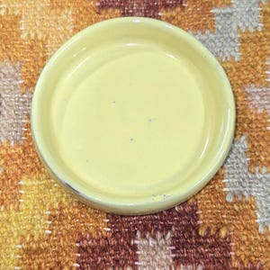 May include: A small, oval-shaped, pale yellow ceramic dish. The dish has a slightly raised rim and a smooth surface. It sits on a colorful, patterned rug with shades of brown, orange, and yellow.