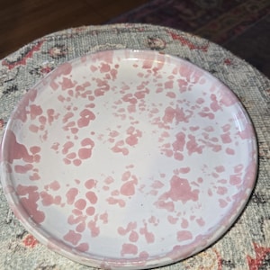 May include: A round, white ceramic plate with a pink speckled pattern. The plate has a slightly raised edge and a glossy finish. The pink spots vary in size and density, creating a unique design. This plate is suitable for serving food.