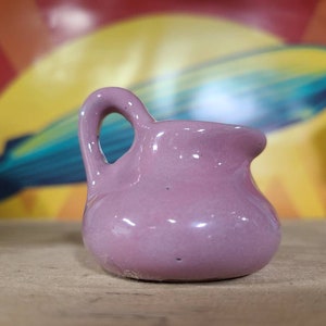 May include: A small, light purple ceramic pitcher with a rounded body and a handle. The pitcher is sitting on a wooden surface.