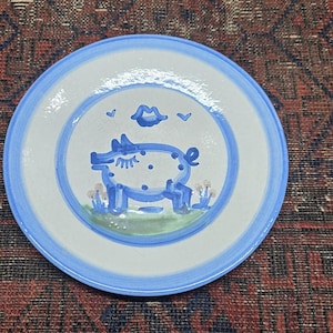 May include: A round ceramic plate with a light gray center and a blue rim. The center features a blue hand-painted illustration of a pig with closed eyes, surrounded by small blue hearts, a pair of lips, and green grass accents at the base.