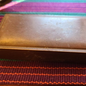 May include: A rectangular, brown box with a slightly raised lid. The box has a matte finish and appears to be made of a solid material. The box is sitting on a colorful striped surface.