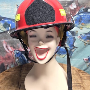 May include: A red firefighter helmet with a black patch and yellow reflective stripes, resting on a mannequin head. The mannequin has a smiling face with red lips. The background features a colorful painting.