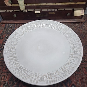 May include: A white ceramic plate with a raised, repeating geometric pattern around the rim. The plate has a smooth, slightly reflective surface and a wide, flat center. The plate is likely for serving food.