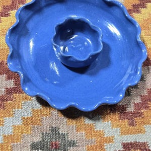 May include: A blue ceramic chip and dip serving set. The set includes a large, round platter with a wavy edge and a smaller, round bowl in the center. The surface has a speckled texture. The set is on a patterned rug.
