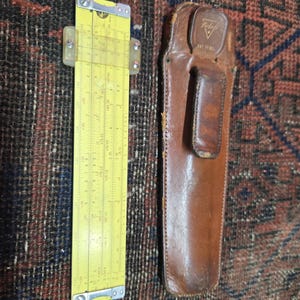 May include: A vintage yellow slide rule with clear plastic guides and metal accents, next to a brown leather case. The slide rule has various scales and markings. The leather case has a belt loop and the text "Pickett Pat. Pend."