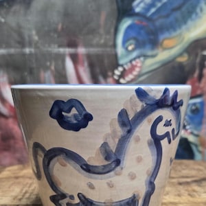 May include: A ceramic cup with a cream-colored base featuring hand-painted blue illustrations of a horse and a pair of lips. The horse design includes light brown polka dots on its body, and the cup has a flared rim with a blue interior edge.