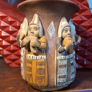 May include: A brown ceramic vase with relief figures of people wearing hats and holding objects. The figures are in brown, tan, and white. The vase has a wide opening and is decorated with geometric patterns.