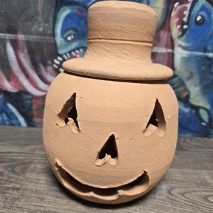 May include: A terracotta pumpkin-shaped candle holder with a top hat lid. The face has triangular cutouts for eyes and a curved mouth. The unglazed ceramic has a light brown color.