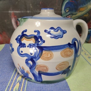May include: A white ceramic pitcher with a blue and brown cow design. The pitcher has a lid and a handle. The cow has brown spots and is surrounded by blue flowers.