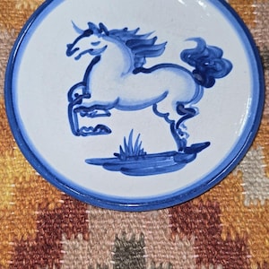 May include: A round, white ceramic plate with a blue rim and a hand-painted blue horse design. The horse is rearing up, with a flowing mane and tail. The plate is on a patterned textile.