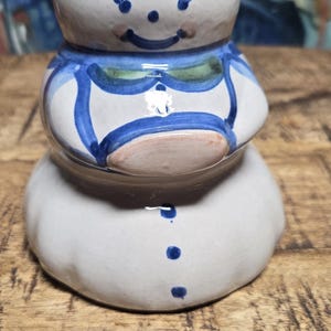 May include: A ceramic figurine shaped like a rounded person with a white base, blue wavy lines, and a painted face. The figure features a green collar, blue accents, and three blue dots down the front. It has a smooth, glossy finish and a rounded top.
