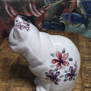 May include: A white ceramic cat figurine with hand-painted floral designs in shades of red, purple, and blue. The cat is in a seated position, with its tail curled. The figurine is on a wooden surface.