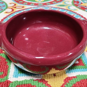 May include: A burgundy-colored ceramic bowl with two small handles. The bowl has a glossy finish and a rounded shape. It is sitting on a colorful patterned surface.
