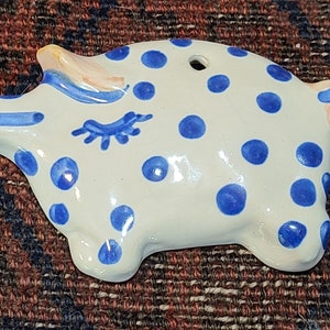 May include: A white ceramic pig ornament with blue polka dots and a blue eye. The pig has a hole in the top for hanging.