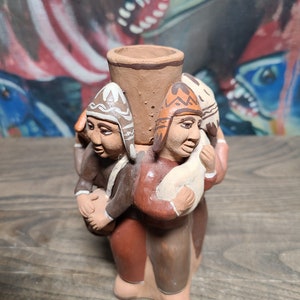 May include: A ceramic figurine of three people, each wearing a hat and holding onto each other. The figurine is brown and has a white base. The figurine is a traditional folk art piece.