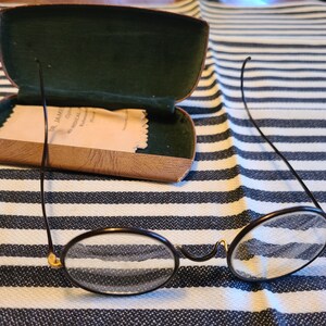 May include: A pair of round, black-framed glasses with gold accents. The glasses are resting on a black and white striped surface. The glasses are in a brown leather case with a green lining.