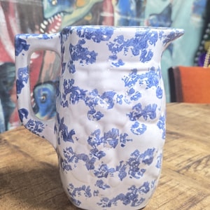 May include: A white ceramic pitcher with a blue floral pattern. The pitcher has a handle and a spout. It is sitting on a wooden surface. The background features a painting of blue fish.