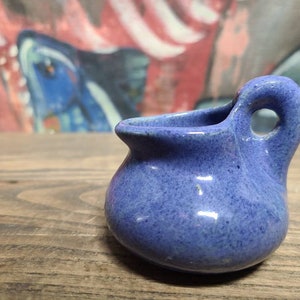 May include: A small, blue ceramic pitcher with a handle. The pitcher has a speckled glaze and a glossy finish.