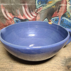 May include: A blue ceramic bowl with a single handle. The bowl has a slight glaze and is slightly chipped on the rim.