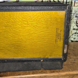 May include: A vintage black box with a textured, golden-yellow panel. The box has a rectangular shape and appears to be made of metal or a similar material. The panel has a unique, wavy pattern. The box is sitting on a wooden surface.