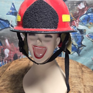 May include: A red firefighter helmet with a black front section and yellow accents. The helmet is on a mannequin head with a smiling face. The background features a colorful painting.