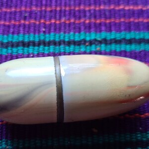 May include: A small, oval-shaped, cream-colored pill box with a gold band around the middle. The box has a swirl pattern in shades of gray, pink, and white.
