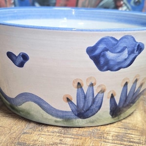 May include: A ceramic bowl with a light beige exterior and a blue rim. The bowl is decorated with blue floral and heart-shaped designs. The base of the bowl has a green stripe.