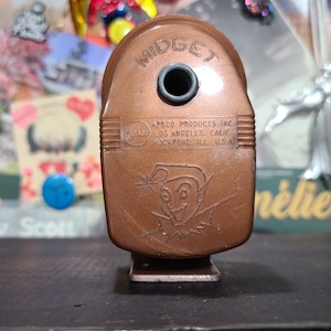 May include: A vintage brown metal pencil sharpener with the word "MIDGET" embossed on the top. The sharpener has a small elf character with a pointed hat and a mischievous grin. The text "APSCO PRODUCTS INC. LOS ANGELES, CALIF ROCKFORD, ILL USA" is printed on the side.