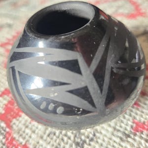 May include: A small, round, black ceramic vessel with a wide opening. The surface is decorated with a light-colored, geometric design. The vessel is likely handmade and has a smooth, polished finish.