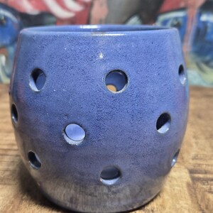 May include: A blue ceramic candle holder with multiple circular cutouts. The holder has a textured surface and sits on a wooden surface. The background features a blurred painting with blue and red tones.
