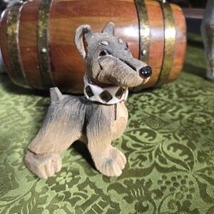 May include: A small, detailed figurine of a dog with gray and tan fur, wearing a white collar with green diamond accents. The dog has a black nose and is standing on a green patterned surface. A wooden barrel is in the background.