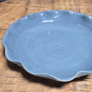 May include: A round, light blue ceramic serving platter with a ruffled edge. The plate is centered on a rustic, wooden table. The platter's color is a muted, dusty blue, and it has a glossy finish.