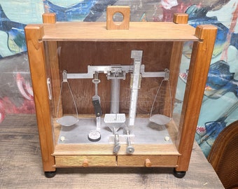 Puede incluir: Una balanza de equilibrio de madera vintage con una caja de vidrio. La balanza tiene dos platillos de metal y una escala graduada para medir el peso. La balanza está en buen estado y tiene un diseño clásico.