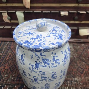 May include: A ceramic jar with a lid, featuring a white base and a blue splatter pattern. The jar has a rounded shape with a matching lid and a small knob. The background includes a wooden cabinet.