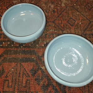 May include: Two light blue ceramic bowls, one slightly larger than the other, on a red and black patterned rug.