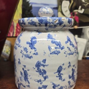 May include: A white ceramic jar with a lid, adorned with blue splattered patterns. The jar has a rounded body and a small, spherical knob on the lid. The design gives it a rustic, handcrafted appearance.