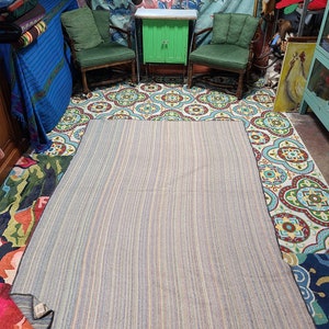 May include: A striped, multi-colored rug with a dark brown border. The rug is laid out on a patterned tile floor with two green chairs and a green cabinet in the background.