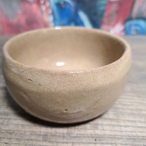 May include: A small, light brown ceramic bowl with a slightly rounded shape. The bowl has a smooth, slightly textured surface.