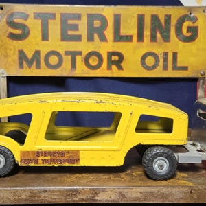 May include: A vintage yellow metal car carrier toy truck with a silver cab and black wheels. The trailer features a yellow frame with open sides and a weathered appearance. Behind the truck is a rectangular yellow sign reading STERLING MOTOR OIL.