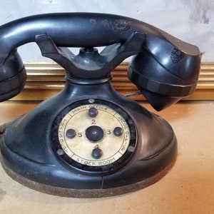 May include: A black rotary dial phone with a cream-colored dial face. The dial face has the text "Main Office" and "Stock Room" printed on it. The phone has a black cord attached to it.