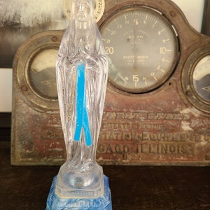 May include: A clear plastic statue of the Virgin Mary with a blue ribbon and a gold halo. The base is blue and white. The statue is in front of an antique gauge.